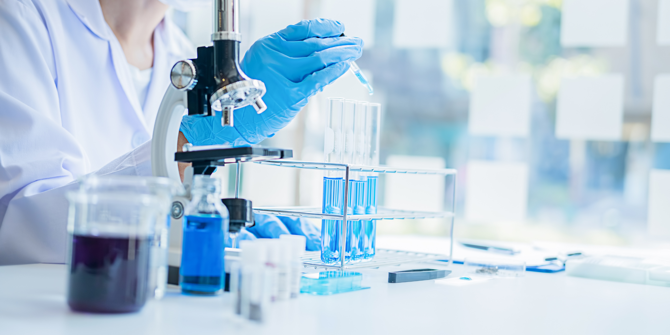 A researcher placing a liquid sample into a test tube, with a beaker of other liquid and a microscope nearby.